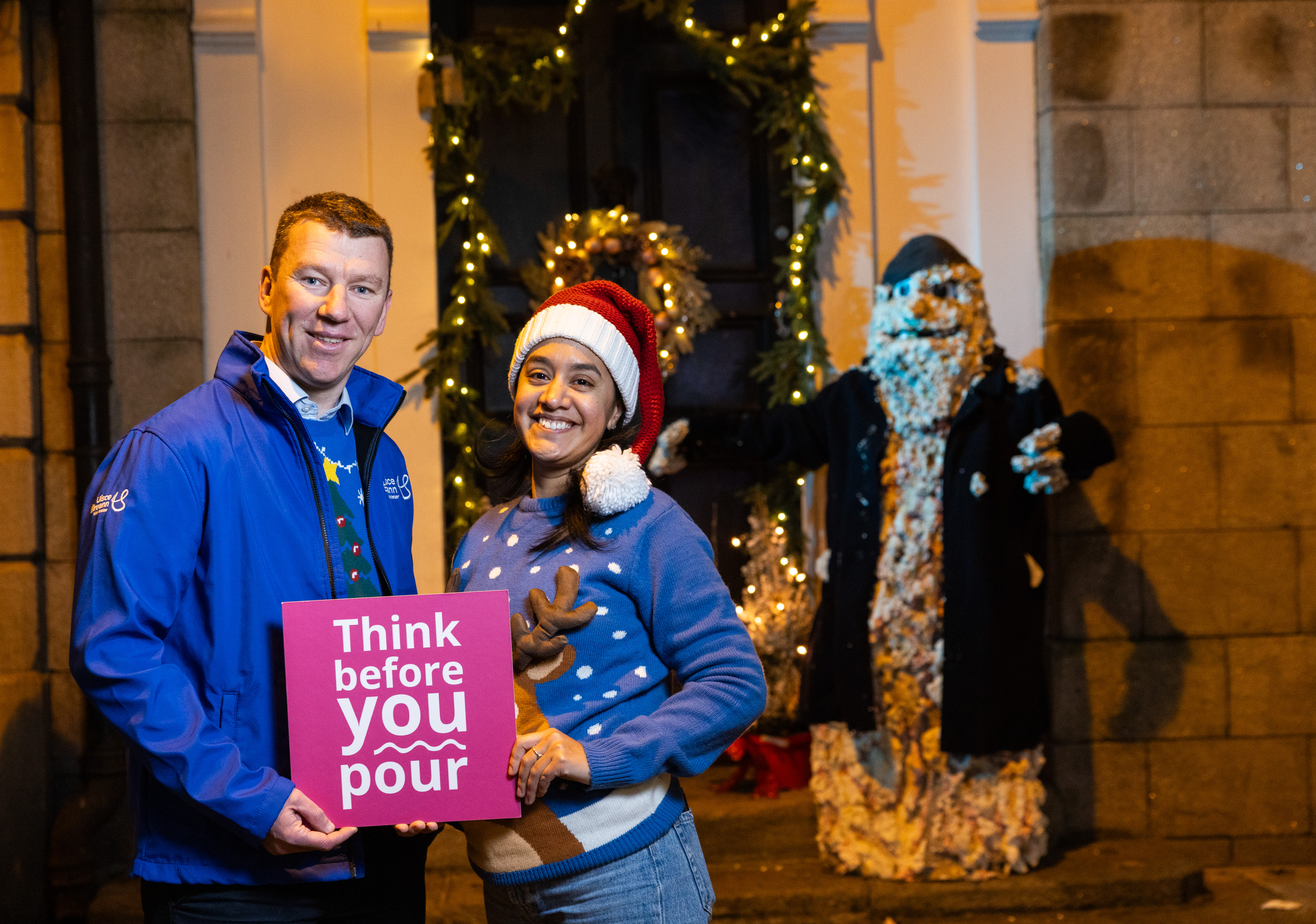 Two people in Christmas jumpers holding a sign reading "Think before you pour"