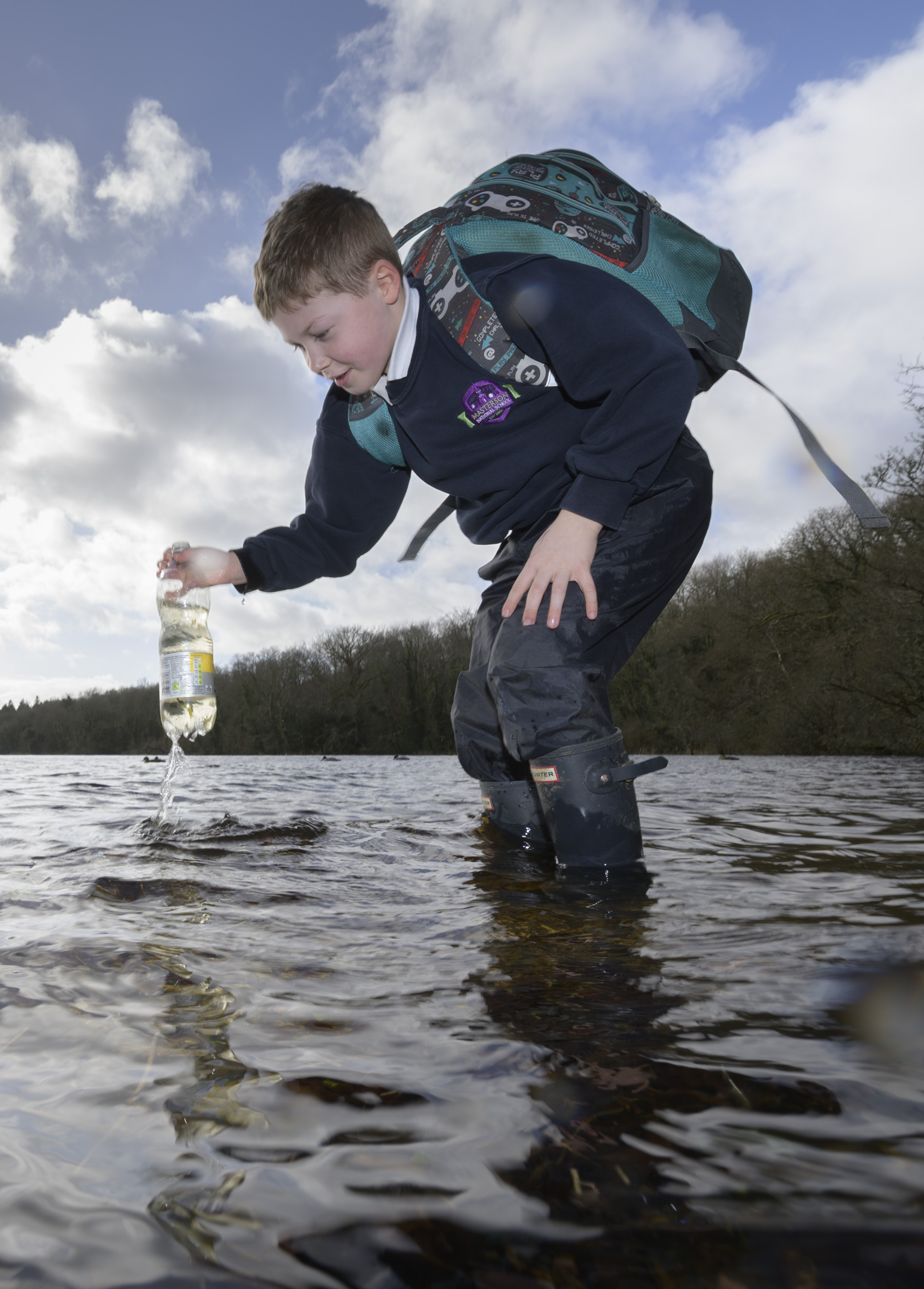 A boy in uniform and wellies with a large schoolbag holding a plastic water bottle over a river he is standing in 