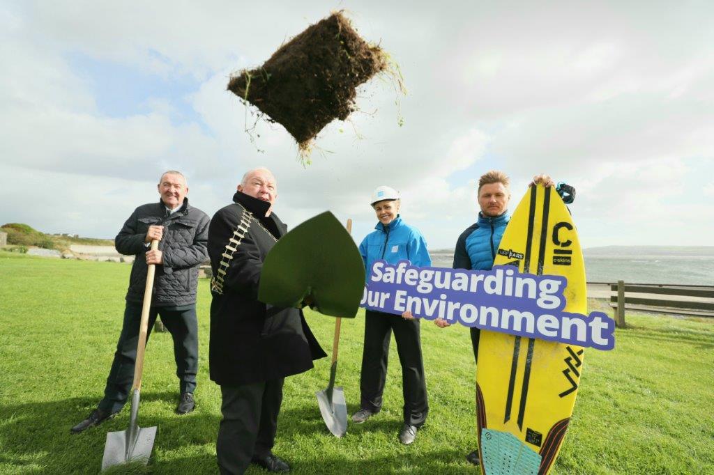 Three people holding shovels, and one man holding a surfboard with a sign reading "Safeguarding our environmant"