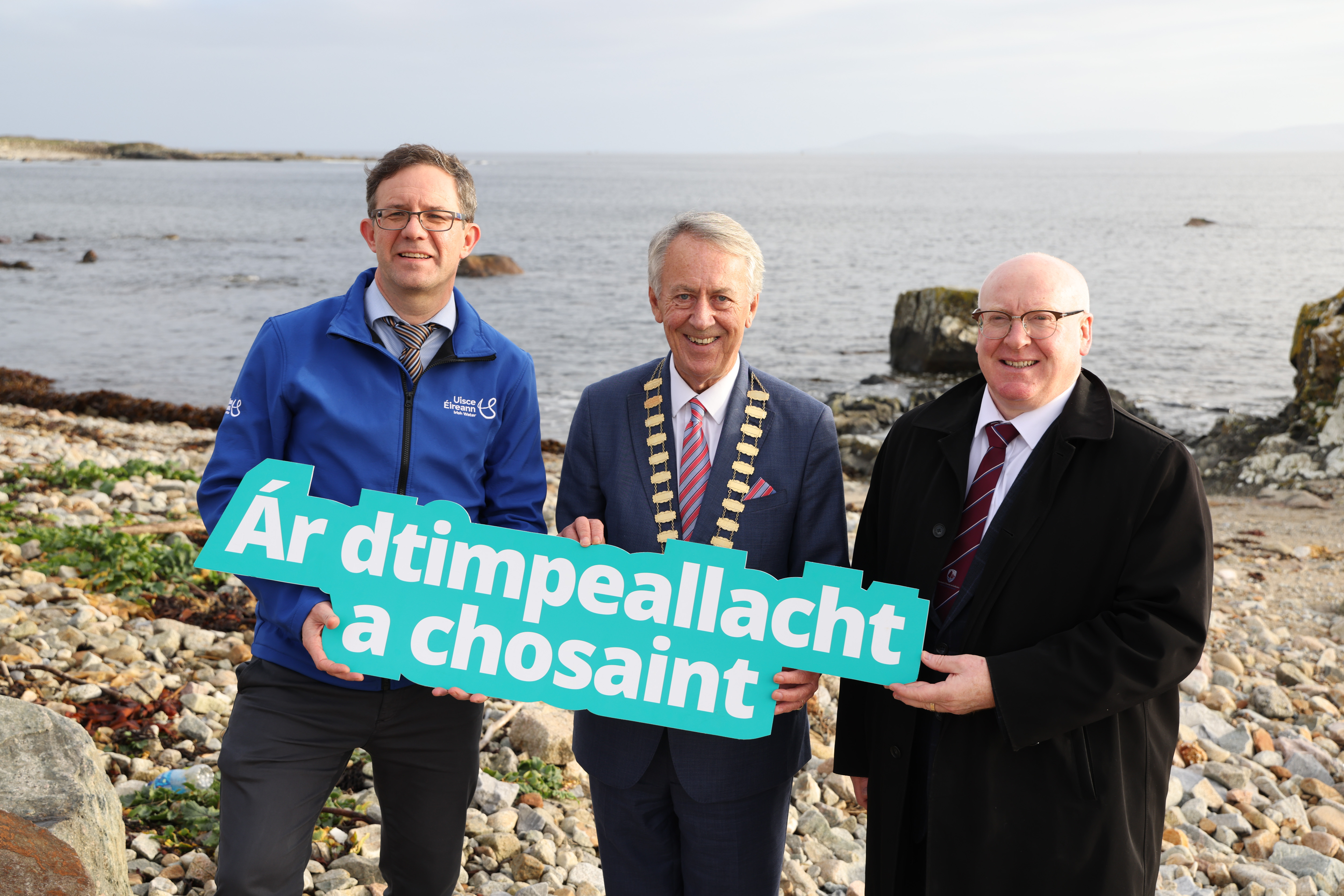 Three men holding a sign reading "Ár dtimpeallacht a chosaint" on a beach